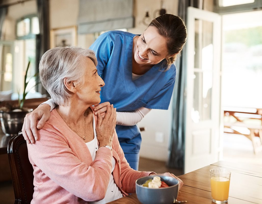 An elderly woman sitting at a table, smiling and looking up at a caregiver who is leaning over her, smiling warmly. The caregiver is wearing blue scrubs and has her arm around the woman's shoulders. There is a bowl of fruit and a glass of orange juice on the table. The setting appears to be a bright, welcoming assisted living or rehabilitation center with open doors and natural light.
