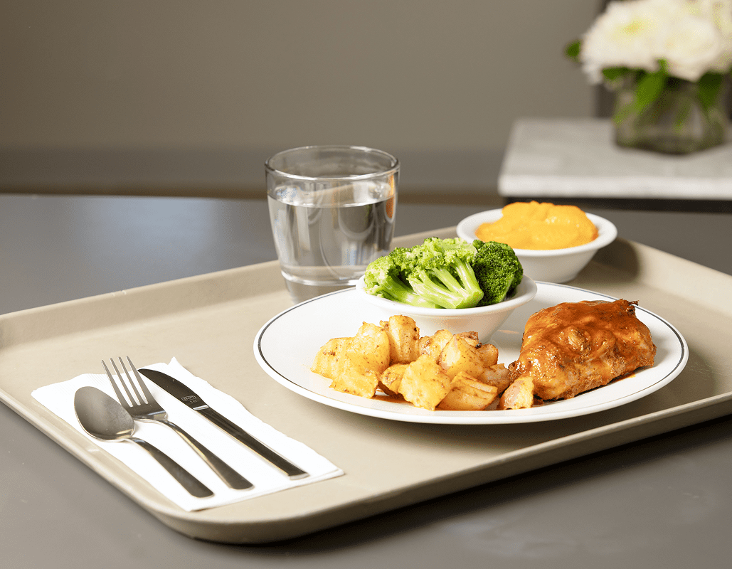 A tray with a meal consisting of broccoli, roasted potatoes, and a piece of chicken with sauce, a glass of water, and a small bowl of mashed sweet potatoes. There are utensils on a napkin on the tray. In the background, there is a vase with white flowers.